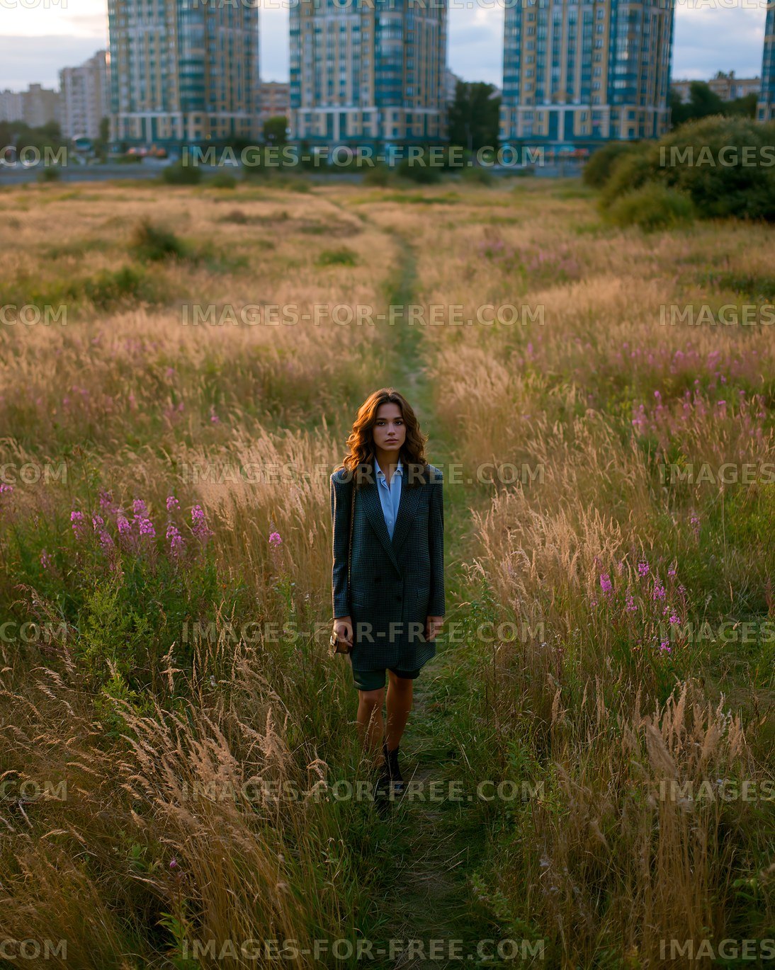 Woman walking on path through tall grass near buildings