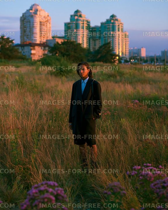 Model standing in a field surrounded by tall grass and buildings